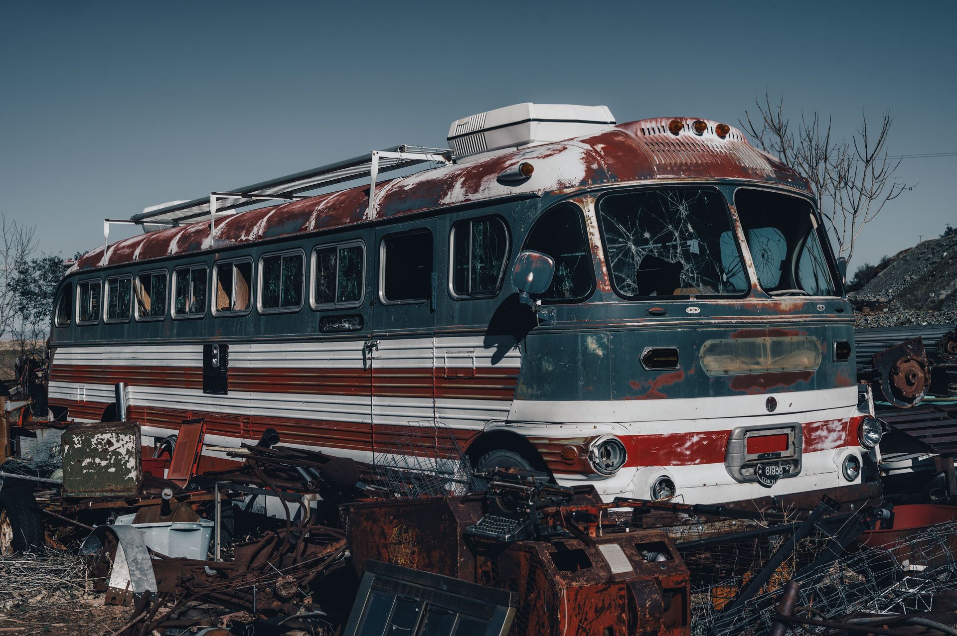 A rusty, abandoned vintage bus parked in a rural junkyard, surrounded by debris under a clear blue sky.