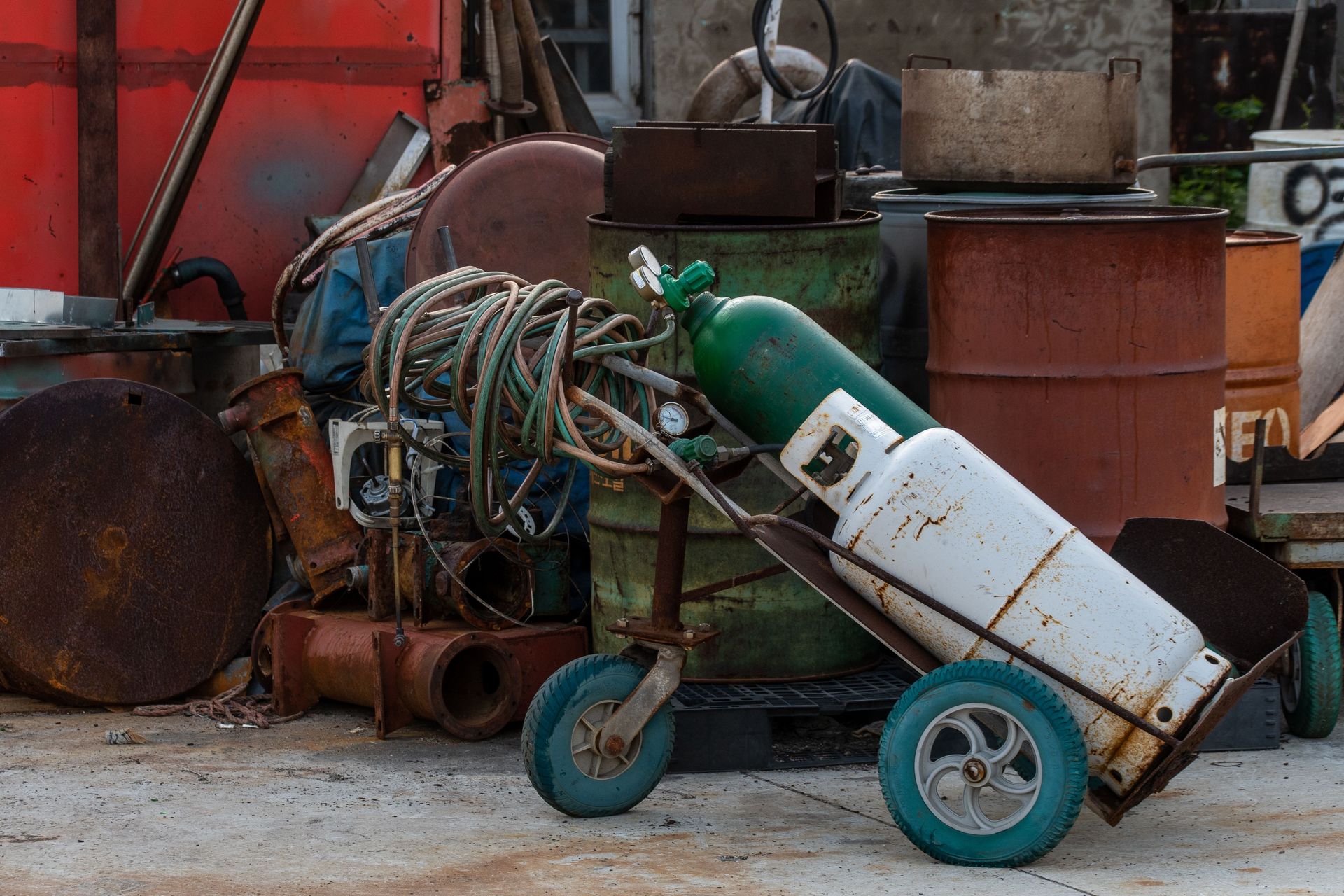 A cluttered industrial junkyard featuring gas cylinders, rusty drums, and tangled hoses.