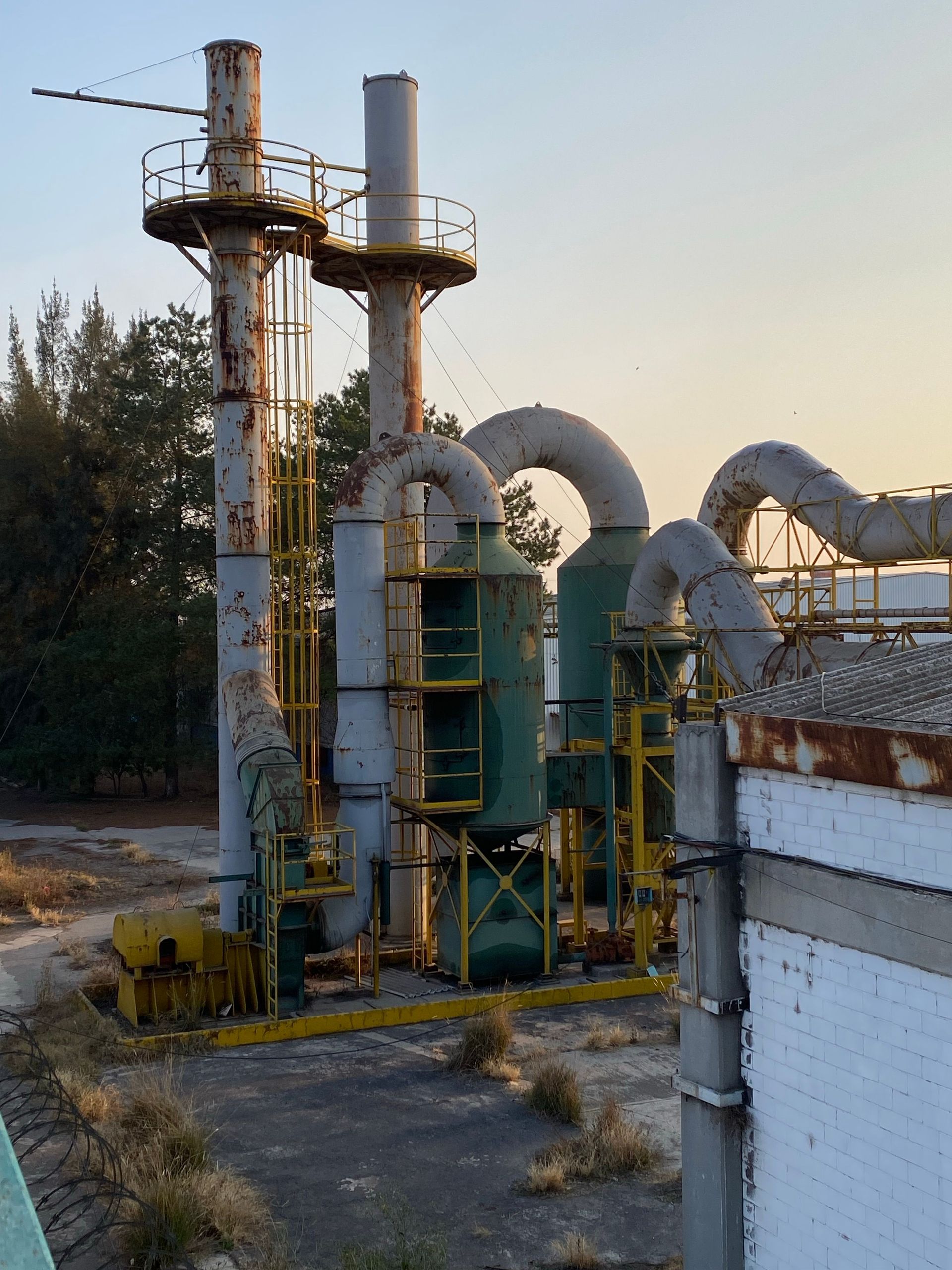 Rusty industrial pipes and machinery outside a factory during sunset, with trees in the background.